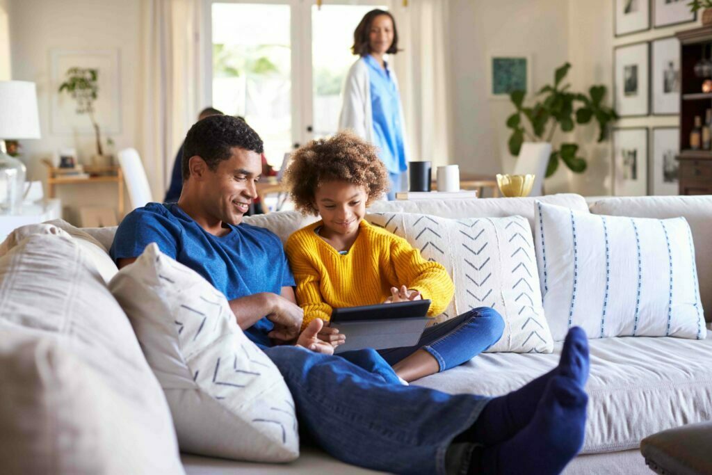 Father and son sit on a couch looking at a tablet with mother in the background.